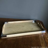 Top angle view of a rectangular blonde wooden serving tray with metal corners and cutout side handles on a brown surface.