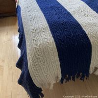 Front view of the throw spread over a surface showing alternating navy and cream vertical cable knit stripes with fringed edges.