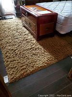 Deep camel shag-style area rug placed on wooden flooring beside a bed and wooden chest, showing thick and plush texture with shag pile.