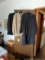 Full view of three men's garments hung on door: black long coat, beige suit jacket, black suit jacket.