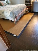 View of natural tan jute carpet with black border extending under bed, measuring approximately 8'x8'. Shows woven texture and condition needing cleaning.