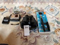 Photo of lot showing Leica Digilux 4.3 camera with battery charger, Tasco binoculars, and Skechers cell phone stick on patterned rug.