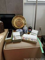 Full lot showing gold metal plate on stand, ceramic dish, bottles, bead doll, mini masks in box, and chopsticks box on chair.