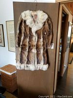 Full front view of muskrat and fox fur coat hanging on a wooden door, showing the length, color pattern, and fur trim.