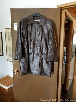 Front view of a brown leather jacket hanging on a door. The jacket is double-breasted with wood buttons, featuring two front flap pockets and a traditional collar.