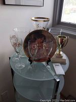 Photo showing four hand-painted Perrier Jouet champagne flutes, three clear glass vases, a gold-tone trophy, and the Knowles display plate on a round glass table near a window.