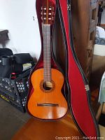 Full view of the Cortez acoustical guitar showing natural wooden finish, nylon strings one of which is broken, and the guitar inside a red-lined black case.