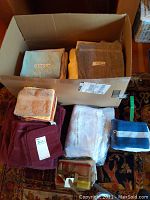 Boxes and bundles of towels in various colors and sizes including grey, beige, brown, maroon, orange, and blue. Different sets and labels on items.
