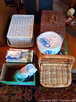 Overall view of three wicker picnic baskets, one white, one natural, and one brown, along with boxes containing disposable cups, cutlery, and plates.