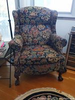 Front view of the floral tapestry wingback recliner showing overall upholstery and carved wooden claw feet