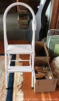 White folding 2-step ladder with curved handle, standing on rug in room