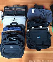 Top-down view of multiple bags including black and blue garment bags, Samboro bag, and canvas bags arranged on wooden floor