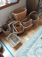 Photo showing various sizes and shapes of woven baskets arranged on wooden floor near a window. Includes rectangular, oval, and round baskets with some having handles.