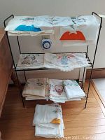 Collection of folded vintage embroidered linens displayed on a metal rack, showing multiple detailed embroidered items.