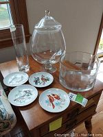 Four bird plates with different bird illustrations and three glass items on a wooden table near a window