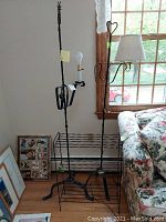 Photo of two metal base floor lamps and one metal shelf placed by a wall next to a window and floral couch, showing lamp details and bases.