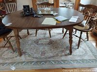 Full view of rectangular wooden dining table with items on top and chairs around it, placed on a floral rug.