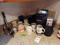 Full view of coffee maker, mugs, accessories, and K-cup box arranged on countertop.
