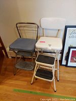 Two vintage Cosco step stools side by side; one white with vinyl seat, one silver with blue vinyl seat; both have two-step design.