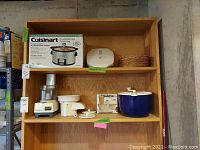 Photo of wooden shelf holding boxed Cuisinart 3 1/2 qt slow cooker, white George Foreman grill, wicker plate holders, small blue salad spinner, and Cuisinart food processor with attachments.