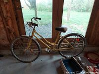 Side profile of the yellow ladies Schwinn vintage bicycle showing frame, seat, and wheels, positioned indoors in front of glass doors.