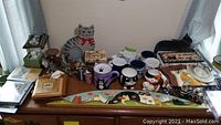 Top-down view of dresser surface with mugs, cat figure, puzzle, puzzle box, wall art panel, and other decor items arranged.