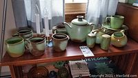 Overview of Francoma Prairie Green pottery pieces including cups, plates, tea pot, sugar bowl, creamer, salt and pepper shakers, and two electric window candles on a wooden shelf.