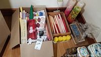 Shows a cardboard box filled with various pastel taper candles, packs of tea lights, and small decorative candles alongside several decorative pillar candles with floral patterns and additional tea lights.