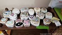 Top view of 16 ceramic music box trinket boxes arranged neatly on a wooden shelf, showing their variety of shapes, sizes and floral/heart themed artwork.