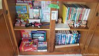 Full view of wooden cabinet shelf containing various board games, VHS movies, children's books, plush toys, and arts and crafts supplies to show overall contents of lot.