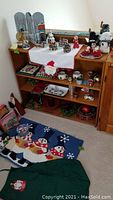 Wooden shelves displaying various Christmas decorations including a signed tole painted Christmas collage, Dickens Heritage Village figures, stuffed toys, Snowfolk figures, mugs, candles, and small holiday decor items.