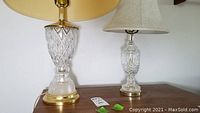 Two crystal lamps on a wooden surface, showing gold and silver-tone bases with beige cloth shades.