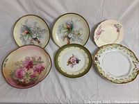 Six hand painted decorative plates including two with bird and blue flower motifs, one with large pink roses, one oval with green rim flower bouquet, and two with pink rose sprays and gold edged detailing.