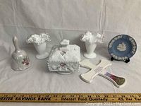Photo shows collection of milk glass and porcelain items including a square covered candy dish, two ruffled edge vases, Wedgwood miniature plate, porcelain bell, and decorative cross arranged on a cloth background with ruler for scale.