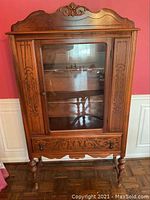 Front view of the antique wood china cabinet against a red wall, showing intricate carvings on the top crest, door panels, and drawer front. The cabinet stands on turned legs with a horizontal stretcher.