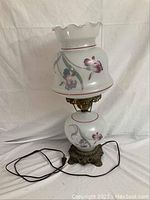 Full view of the glass lamp showing the hand-painted iris floral pattern on white glass shade and base with brass-toned ornate metal base. Black electric cord attached.