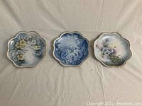 Three decorative blue floral china plates arranged side by side on a light background showing scalloped edges and gold trim.