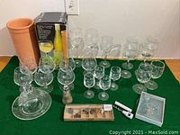 Various wine and brandy glasses, terracotta wine cooler, wine bottle charms, cheese server and other wine accessories arranged on a green cloth.