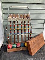 Front view of croquet set shows six wooden mallets with wrapped handles and colorful stripes, six colorful balls, wicket, and wooden rack with metal handle.