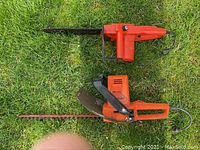 Top-down view of both the red corded chainsaw and the red corded hedge trimmer on grass.