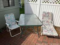 Wide view of outdoor patio set on brick patio including glass topped table, folding chair with floral fabric, and folding chaise lounge with floral cushion.