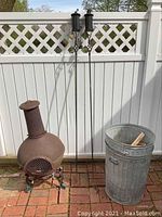 Full view of iron chiminea, two torches, and two galvanized barrels lined against white fence outside on brick ground.