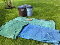 Three Rubbermaid garbage totes with lids, two gray and one brown, with assorted plastic tarps in green and blue spread out on grass.