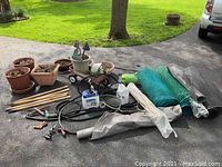 Overview of gardening containers, wooden stakes, hoses, garden fencing rolls, sprayers, and decorative items on driveway.