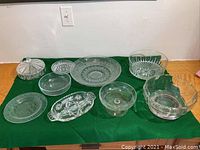 Nine assorted clear glass serving bowls and platters laid out on green cloth, shown from front angle.