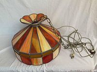 Angle view of the lamp showing the orange, yellow, and cream stained glass panels and the metal chain and cord attached.