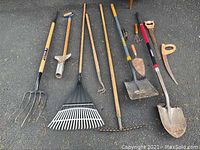 Full view of assortment of long handled yard tools including pitchfork, rakes, hoes, shovels and hand tools spread on pavement surface