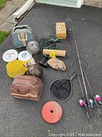Photo showing assortment of sporting goods including fishing rods, tackle box, bowling balls, paddle ball rackets, frisbees, jump rope, dumbbell, and small fishing net on driveway.