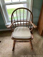 Front view of the wooden Windsor style rocking chair with cushion. Shows the curved back with vertical spindles and seated cushion on the wooden seat frame.