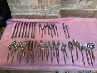 Wide layout of stainless steel flatware on pink cloth, showing mixed types and designs including knives, forks, spoons, and serving utensils.